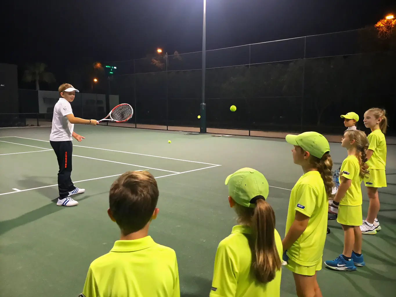 A close-up image of a tennis coach instructing young players during a training session. The coach is demonstrating proper technique, and the players are attentively following along.